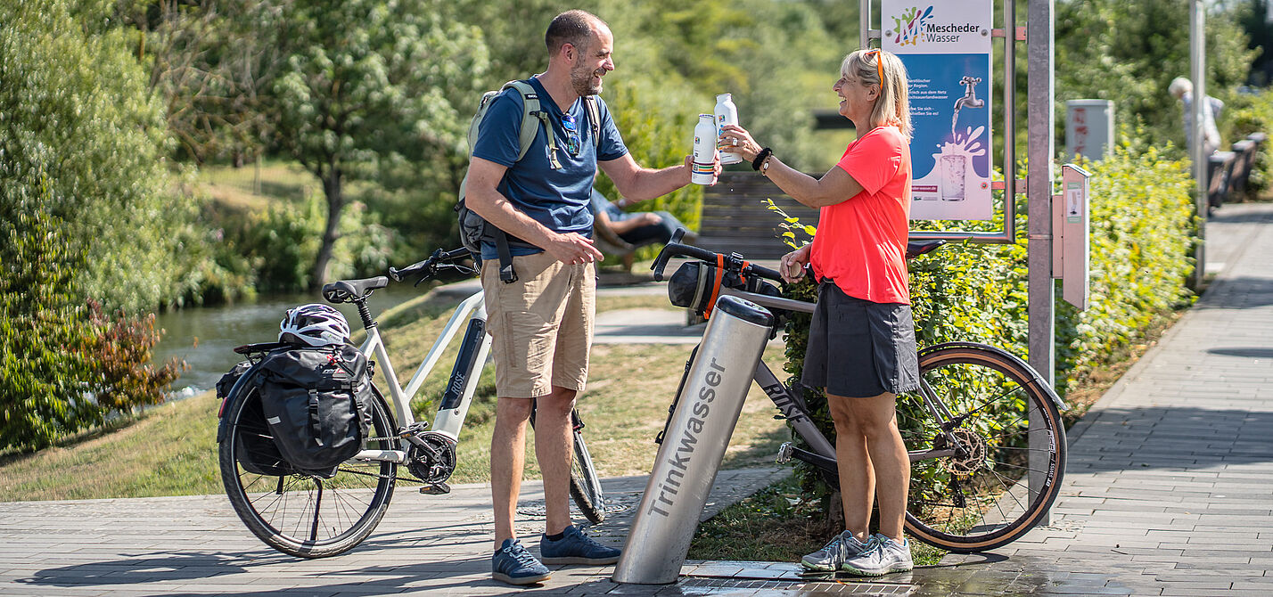 Zwei Radfahrende füllen Trinkflaschen an einem Trinkbrunnen des RuhrtalRadwegs auf.